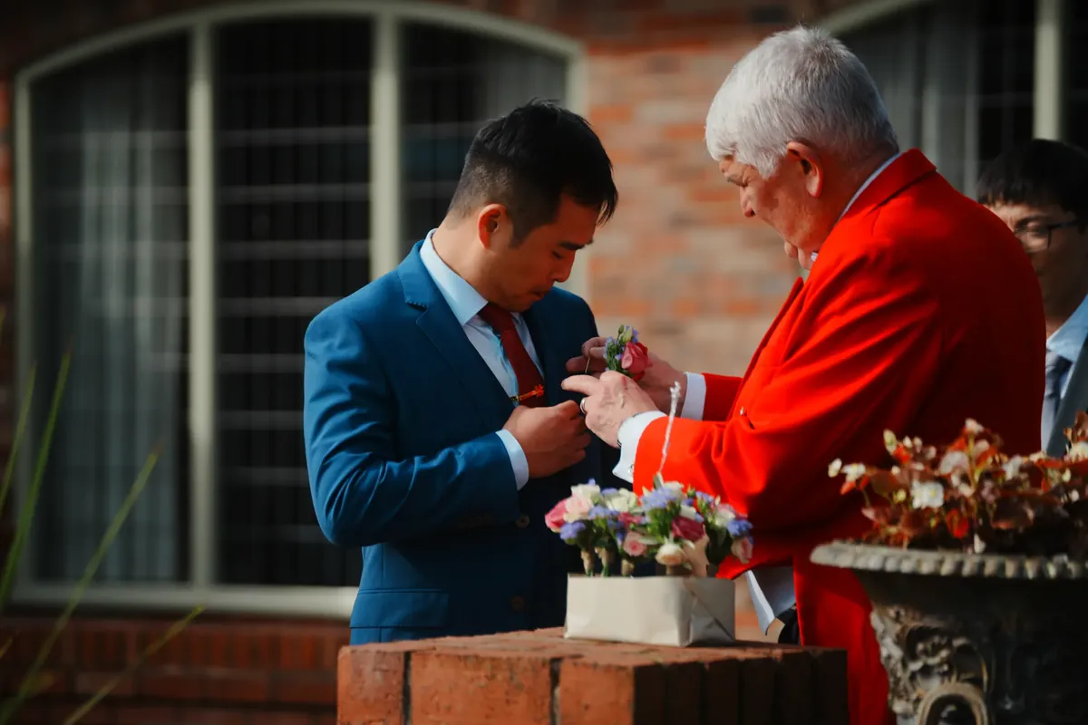 The groom receiving his boutonniere from a red-coated master of ceremonies at Colshaw Hall